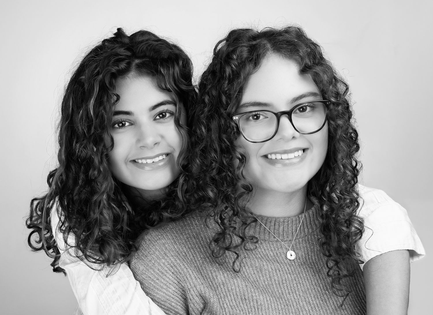 Black and white image of two girls in front of a white/grey backdrop, smiling.  One has her arms wrapped around the other - quarter shot