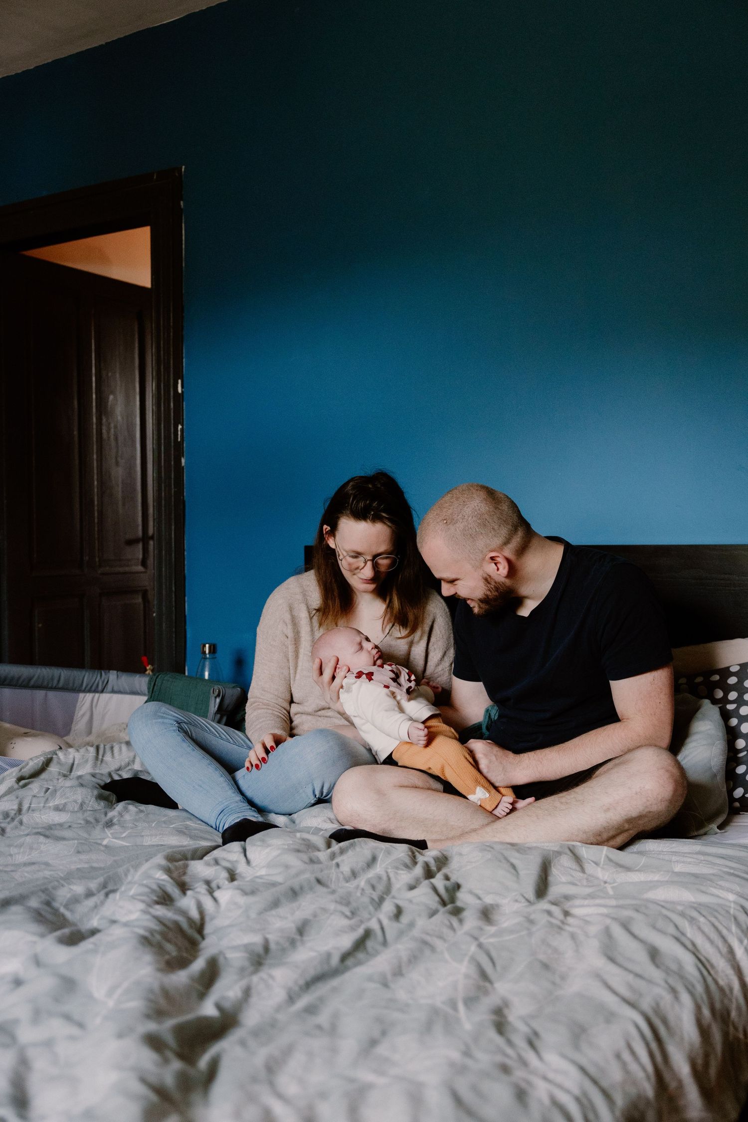 Young parents sitting on a bed with a blue wall, lovingly looking at their newborn baby.