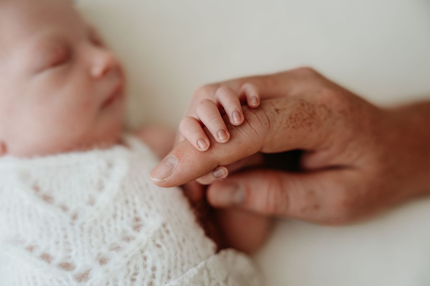 Newborn baby sleeping peacefully while gently gripping an adult finger, wrapped in a white knit blanket.