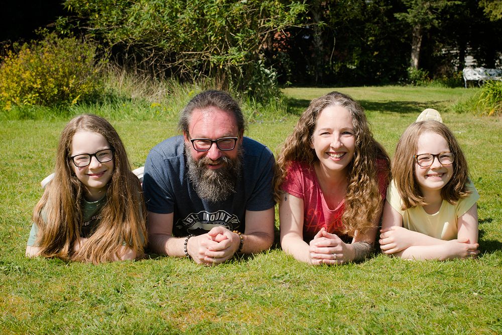 A family of four lies on a sunny lawn, smiling warmly. Two adults are flanked by two children, all wearing glasses. Background features lush