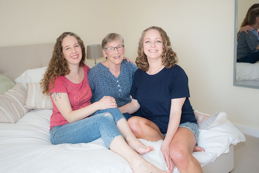Three women sit smiling on a bed in a cozy room. Two younger women flank an older woman in the center, relaxed and happy in casual clothes
