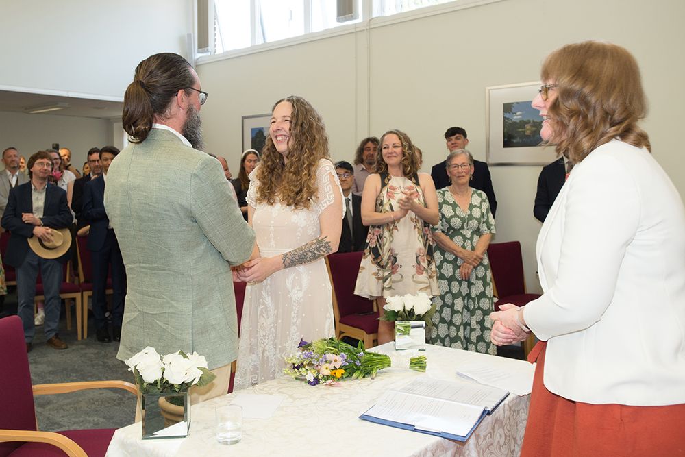 Bride and groom stand smiling and holding hands during a wedding ceremony. Guests, some clapping, and the officiant in white observe joyfuly