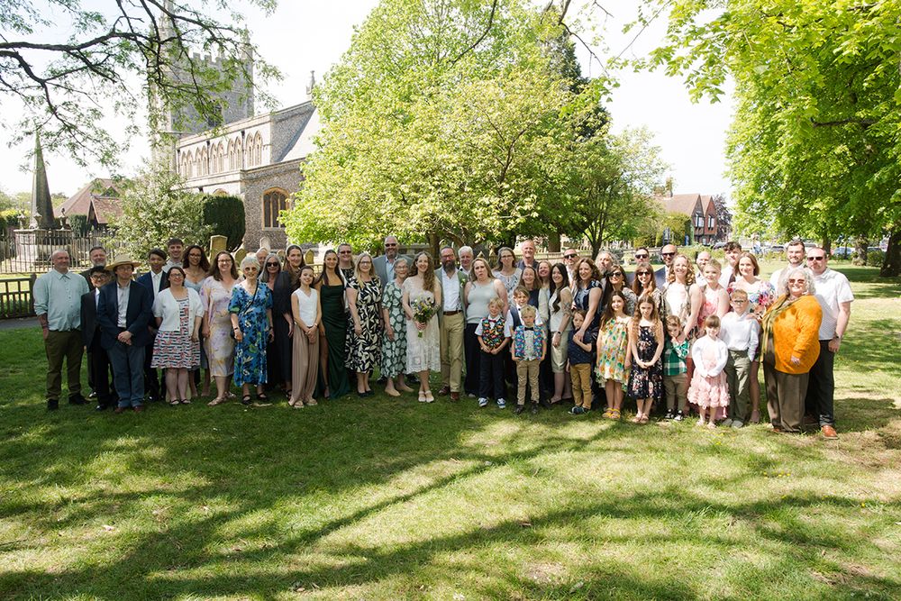 A wedding party poses outdoors on a sunny day in front of a church. They are standing on grass, surrounded by lush green trees.