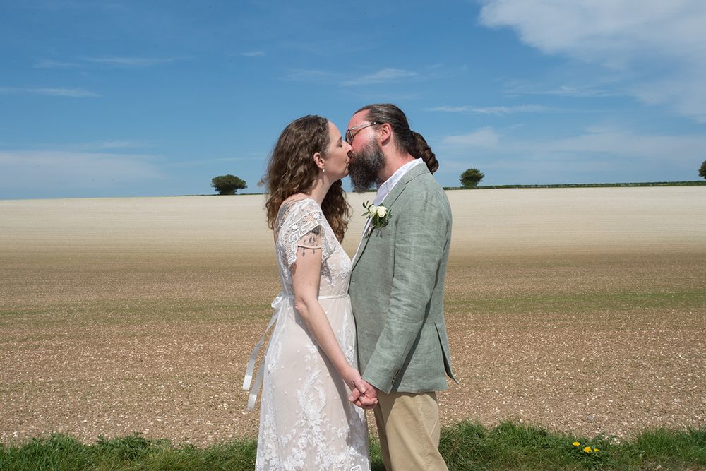 Newlyweds shares a kiss in a sunny field. The woman wears a lace wedding dress, and the man is in a light green jacket. The sky is clear.