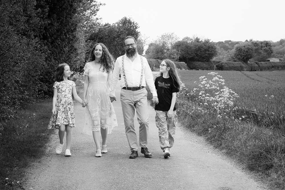 A family of four walks down a country road, holding hands, smiling. The scene is serene, with fields and trees lining the path.