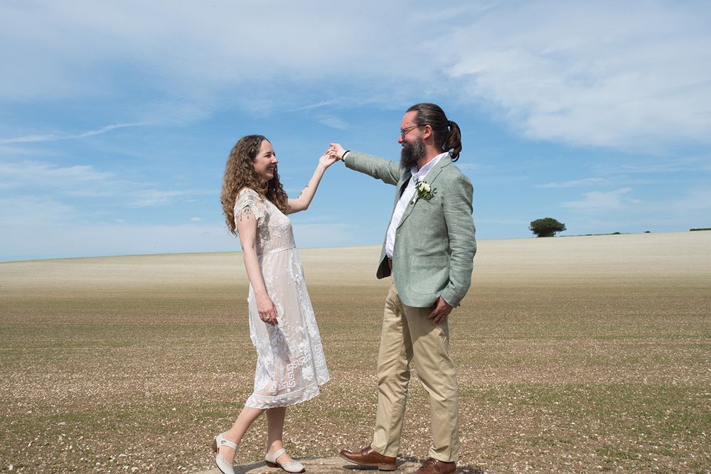 Bride in a white dress and Groom in a green jacket smile while holding hands in a vast field under a blue sky, exuding joy and serenity.