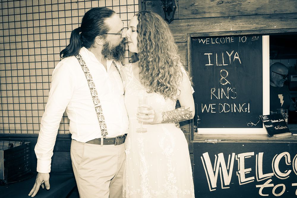 A couple gently touches noses in front of a chalkboard reading "Welcome to Illya & Rin's Wedding." The mood is intimate and joyful.