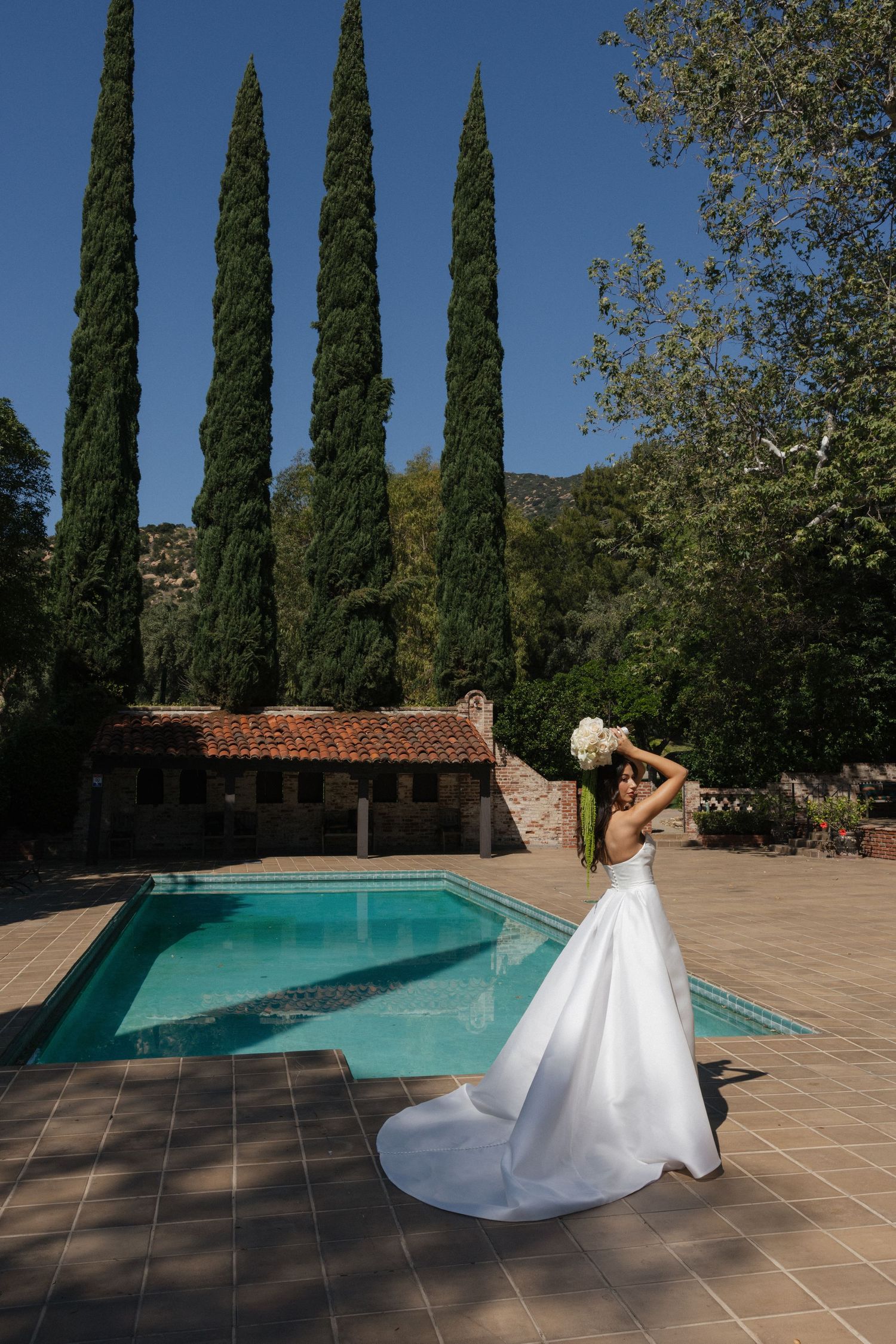 Bride in white gown holding bouquet poses by outdoor pool with cypress trees and terracotta roof building.