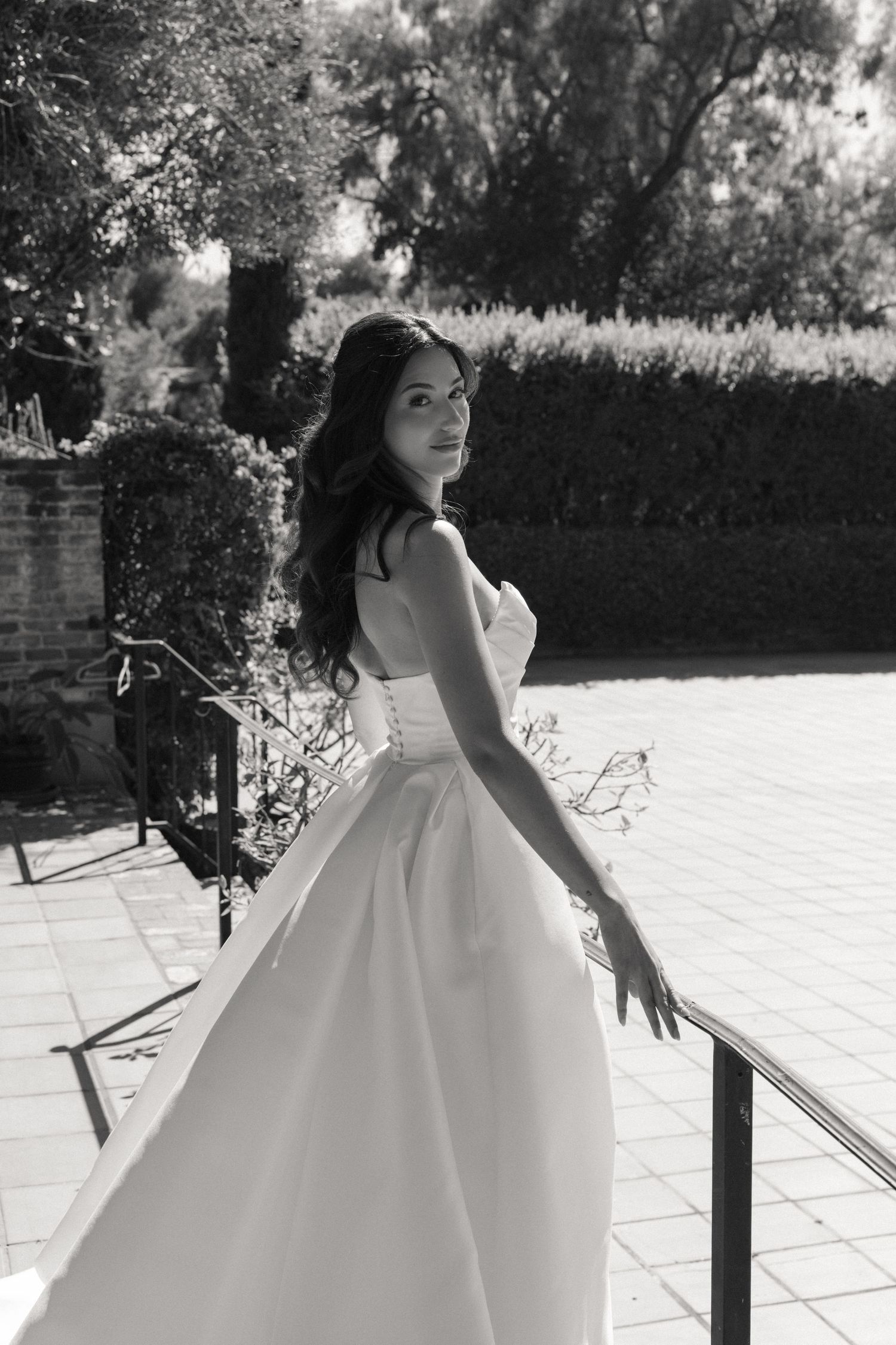 Black and white photo of a bride in a strapless white gown posing on a terrace with lush greenery.