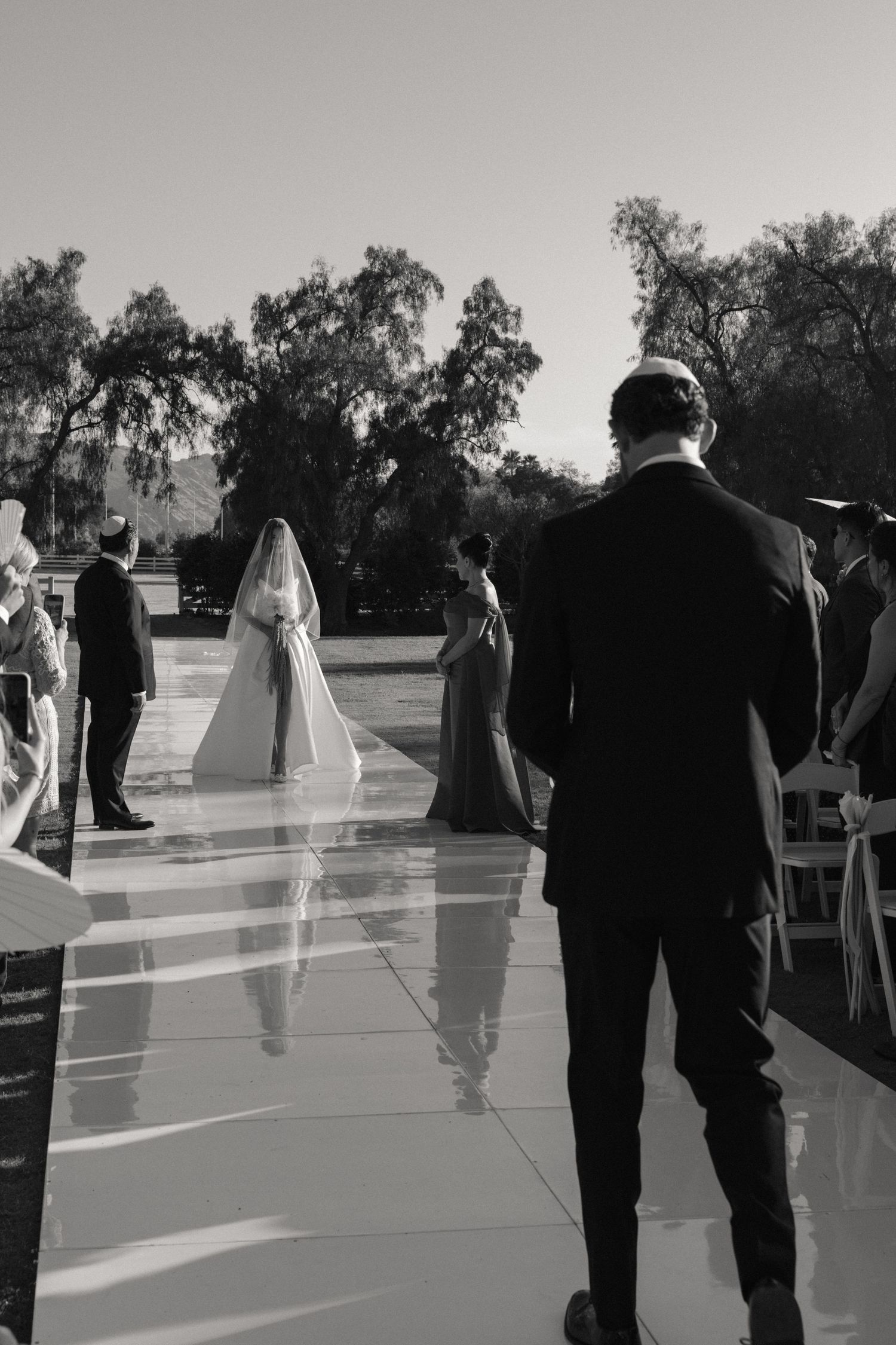 Black and white outdoor wedding ceremony with bride walking down aisle toward groom, trees in background.