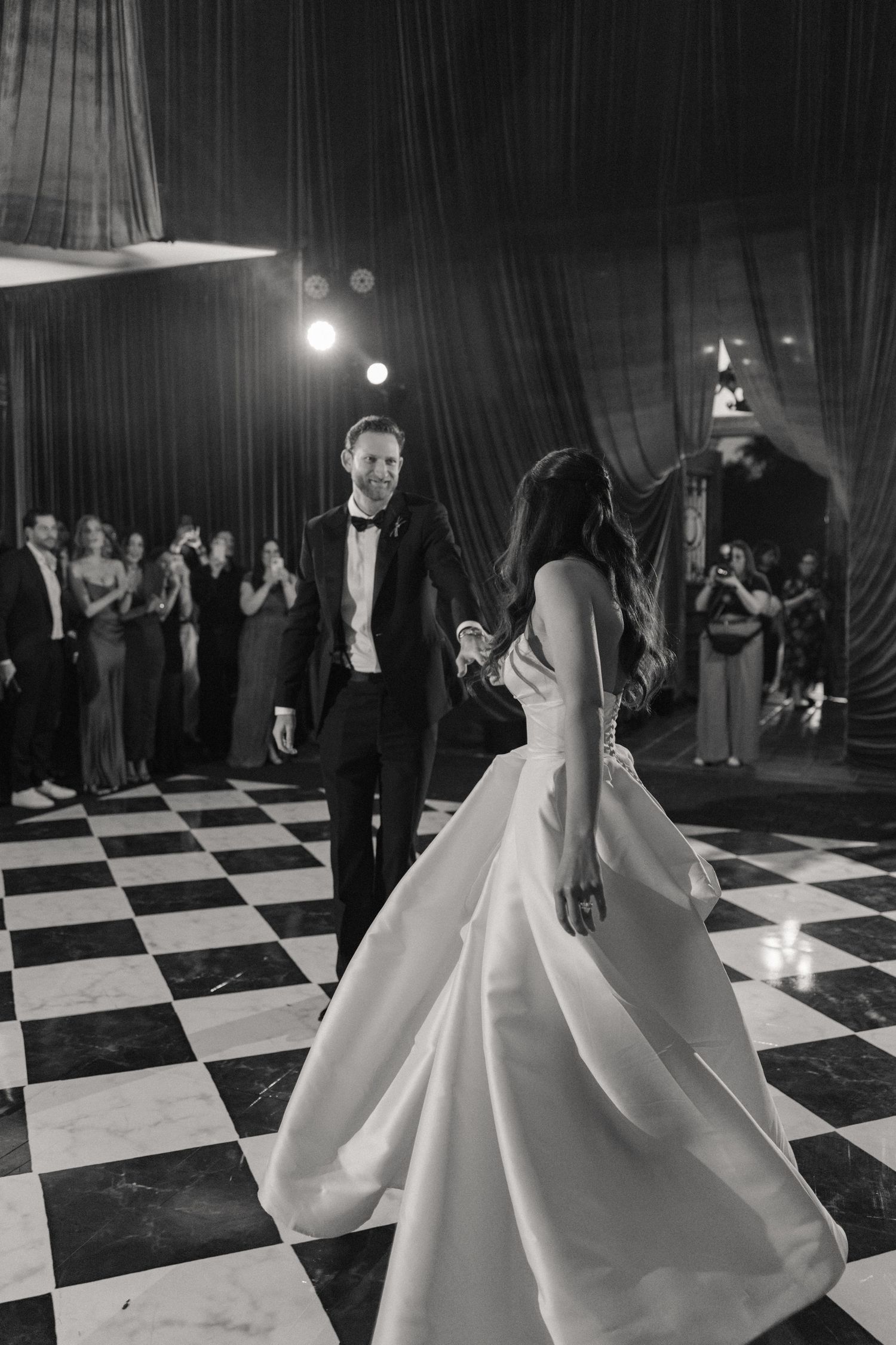 Black and white photo of bride and groom sharing first dance on checkered floor at elegant wedding reception.