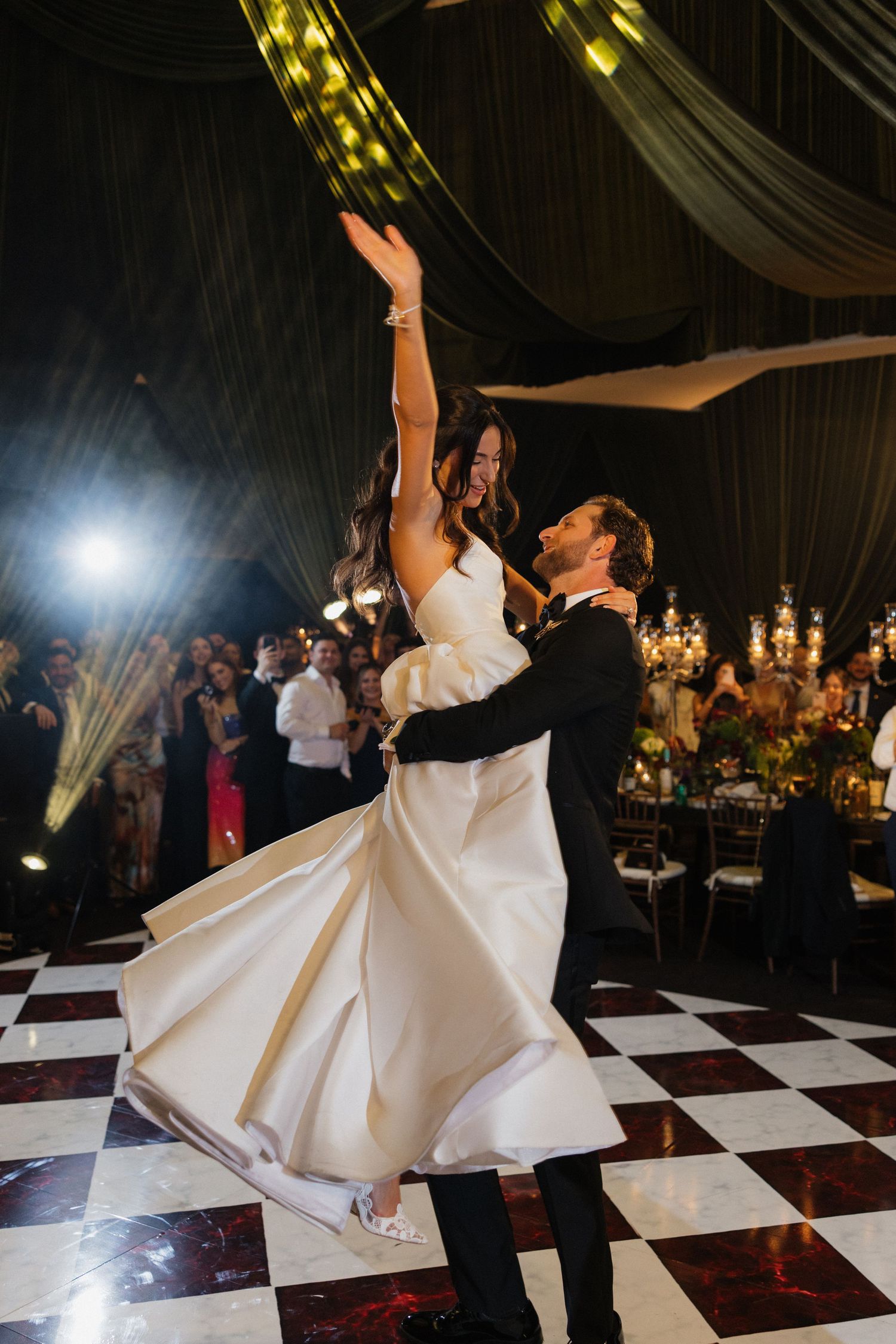 Bride and groom share first dance on checkered floor in elegant ballroom, her white gown swirling as he lifts her arm.