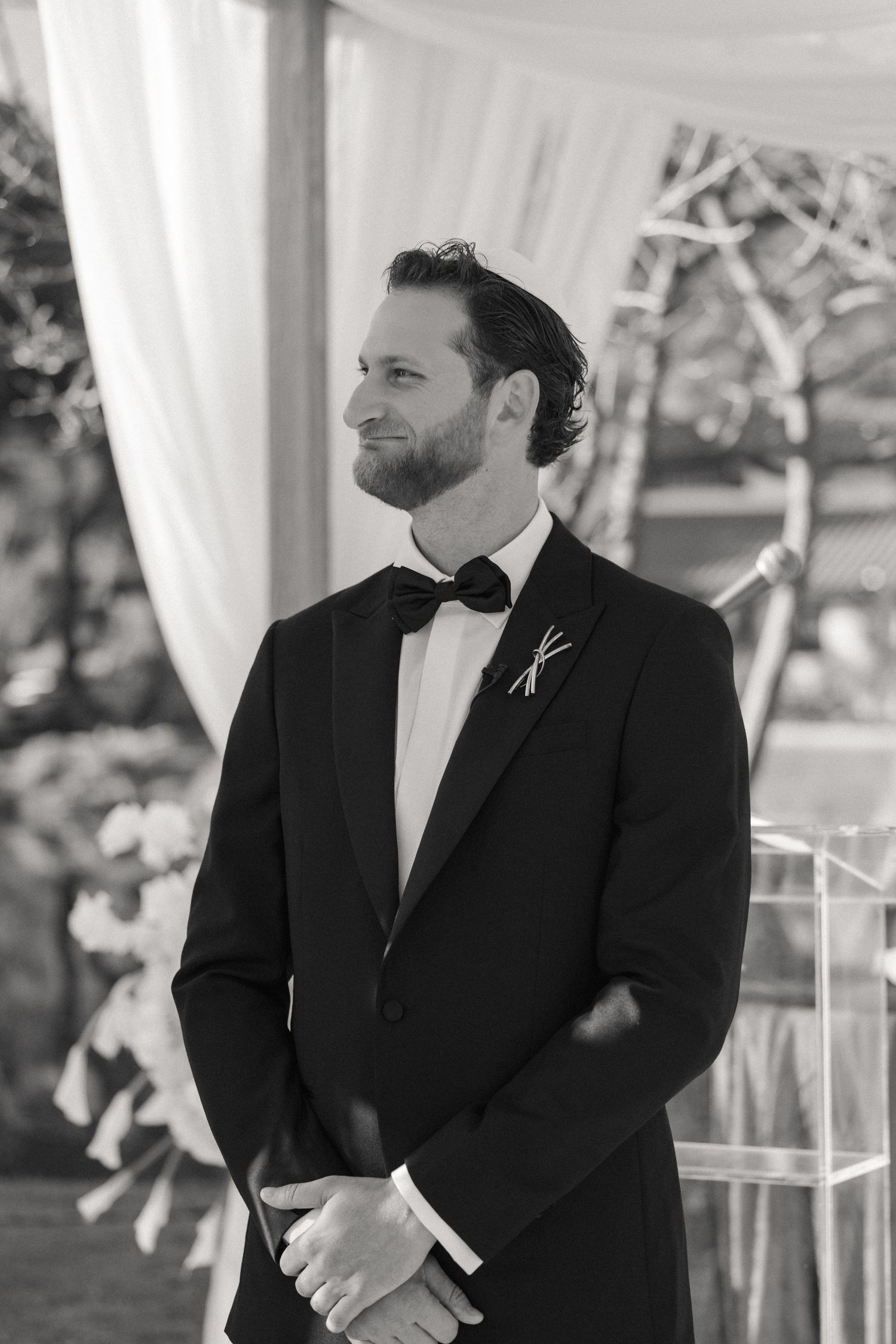 Black and white photo of a groom in a tuxedo with bow tie smiling at the altar during a wedding ceremony.