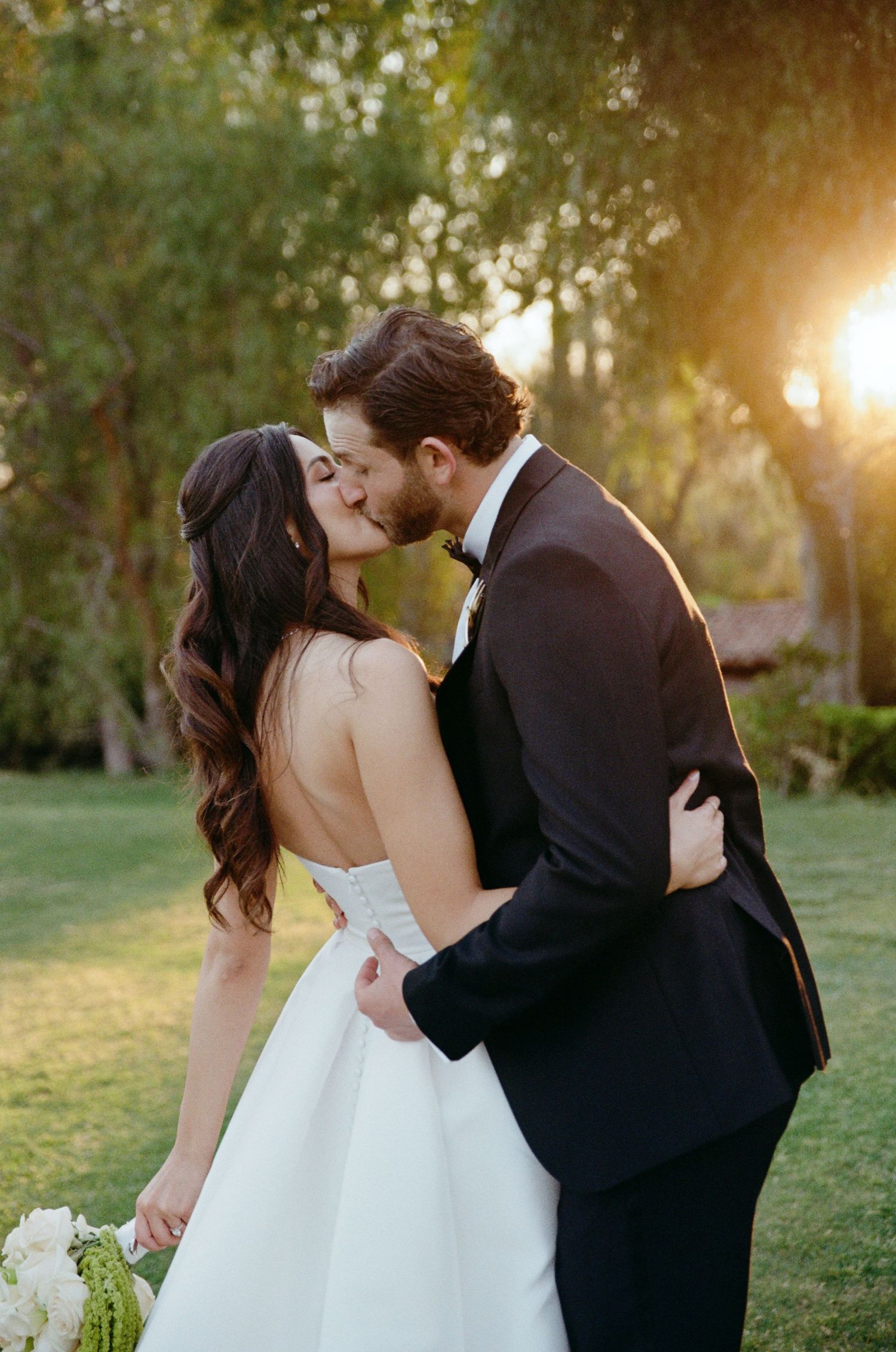 Bride and groom sharing golden hour kiss outdoors, bride holding white bouquet.