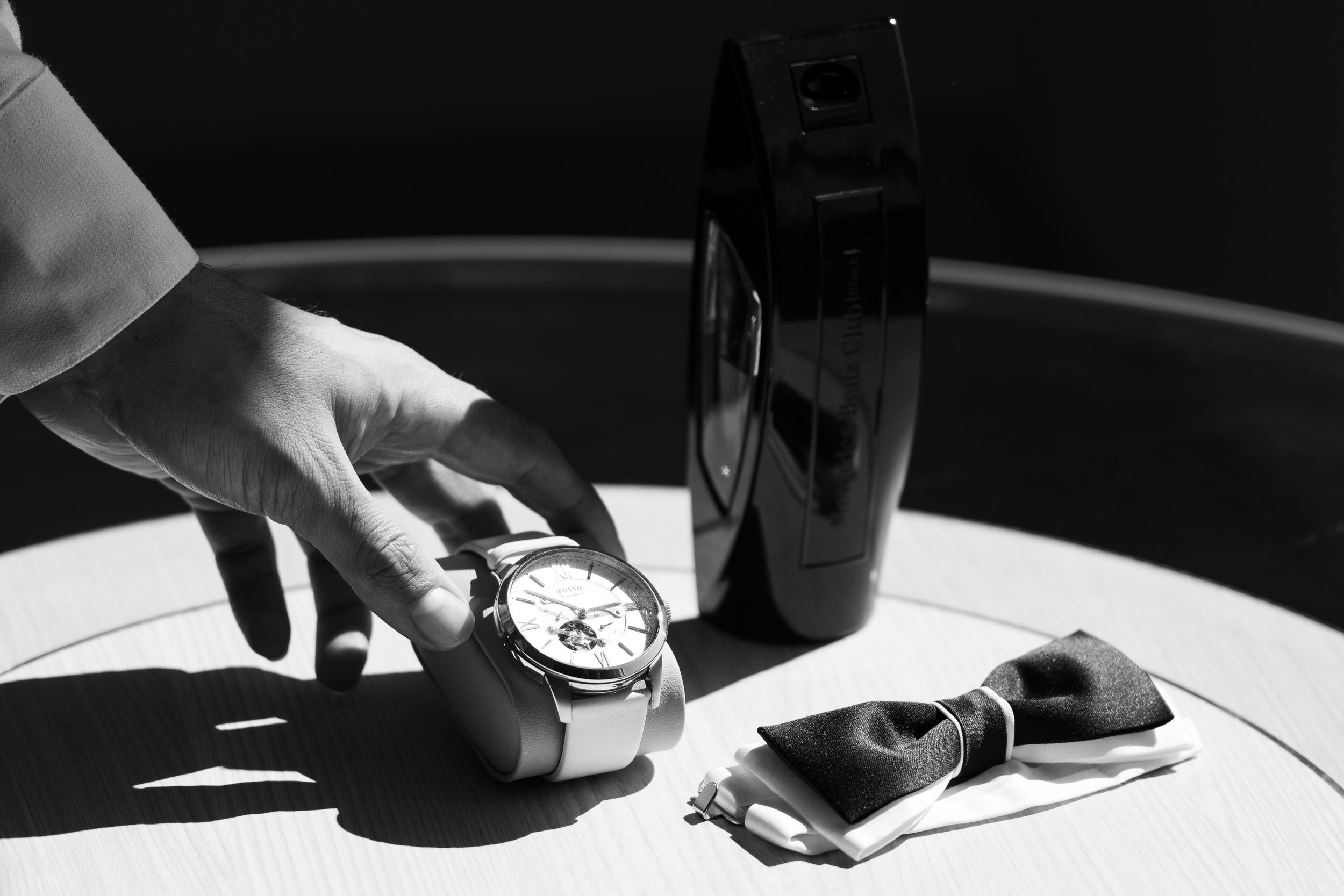 Hand reaching for elegant watch on table beside bow tie and dark vase in black and white luxury setting.