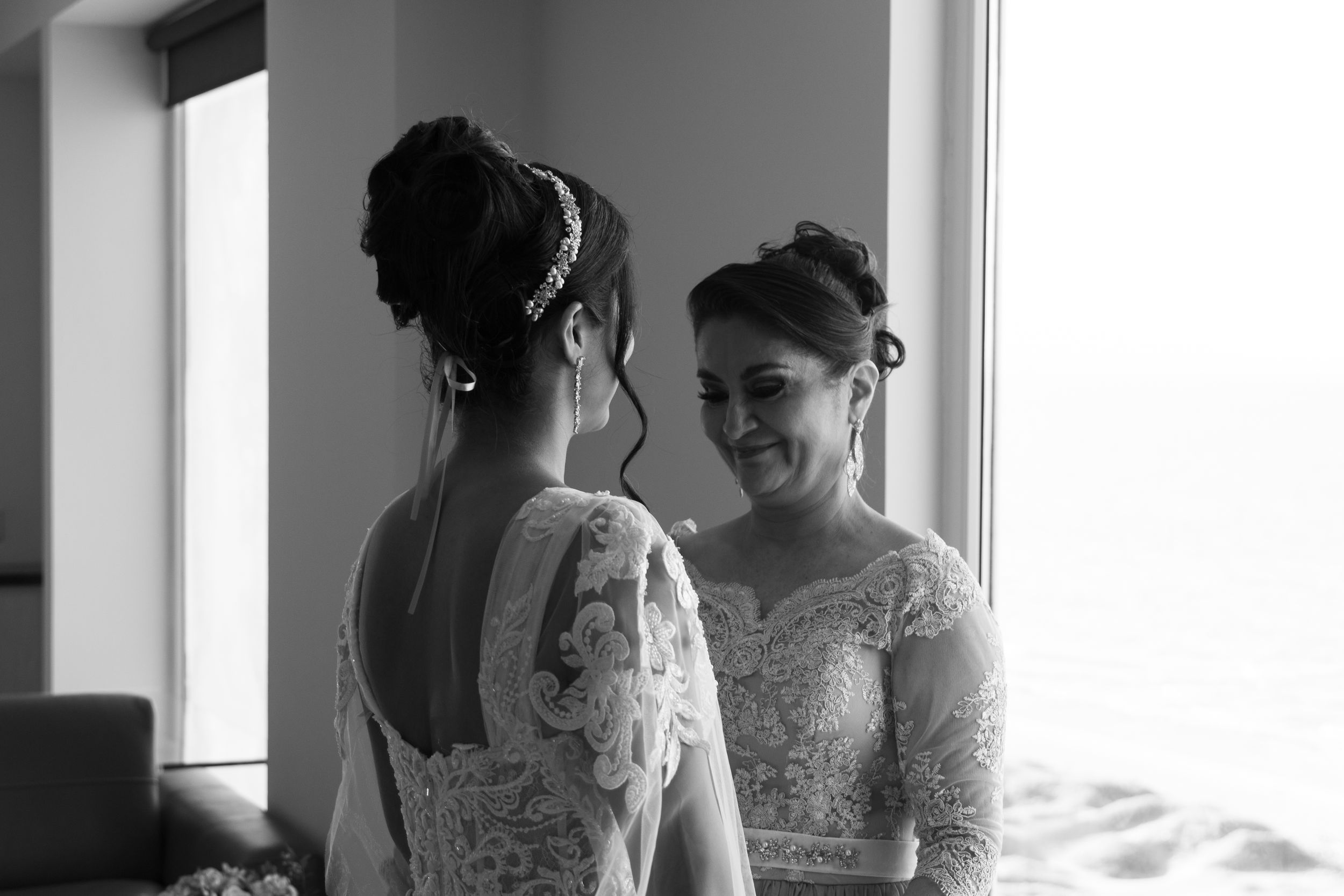 Black and white photo of two brides in lace wedding gowns smiling at each other near a window.