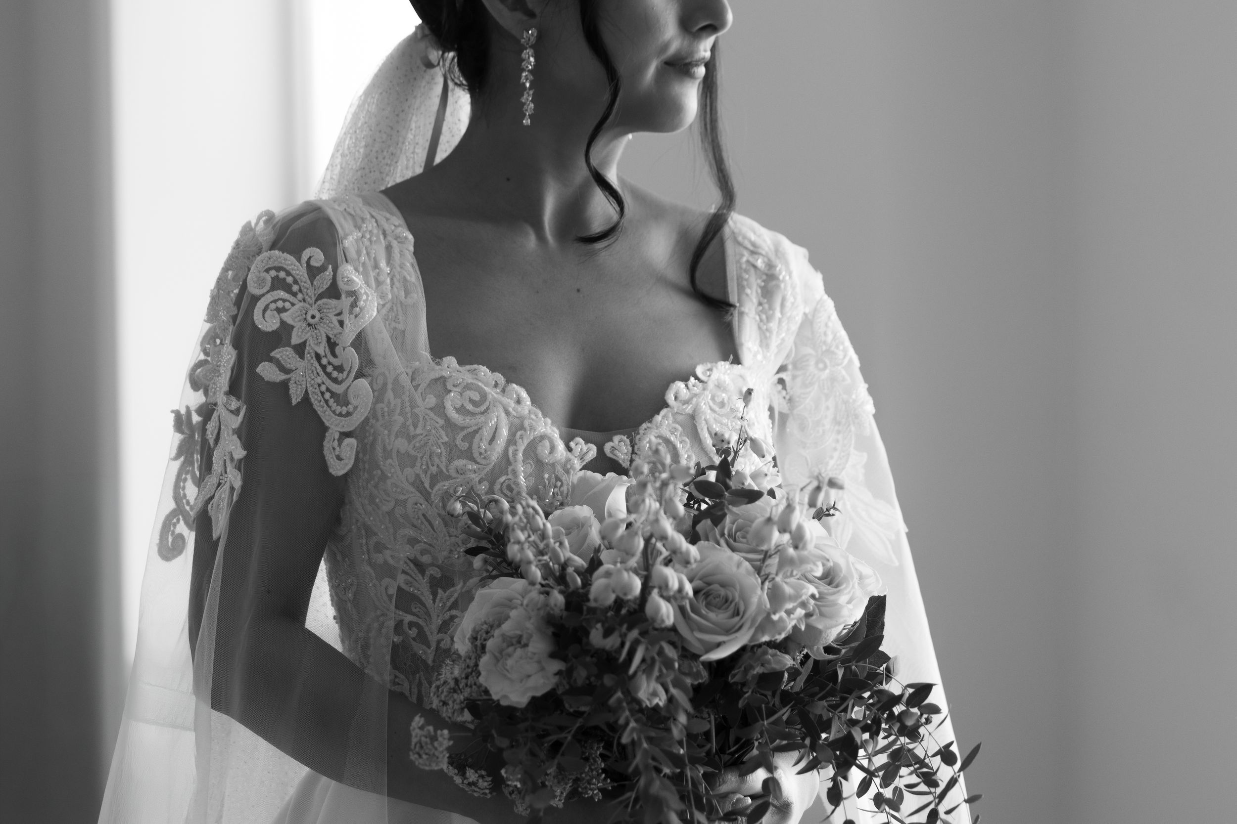 Bride in lace wedding dress holding floral bouquet in elegant black and white portrait.
