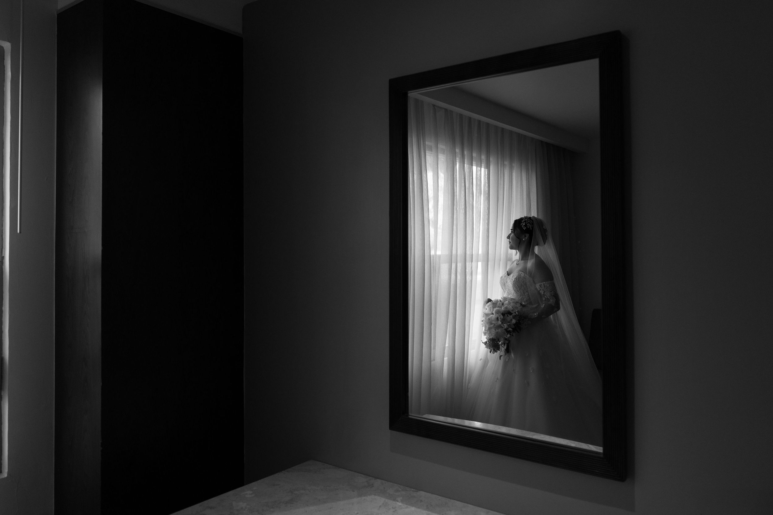 Black and white photo of a bride in a wedding dress reflected in a mirror, holding a bouquet near a bright window.