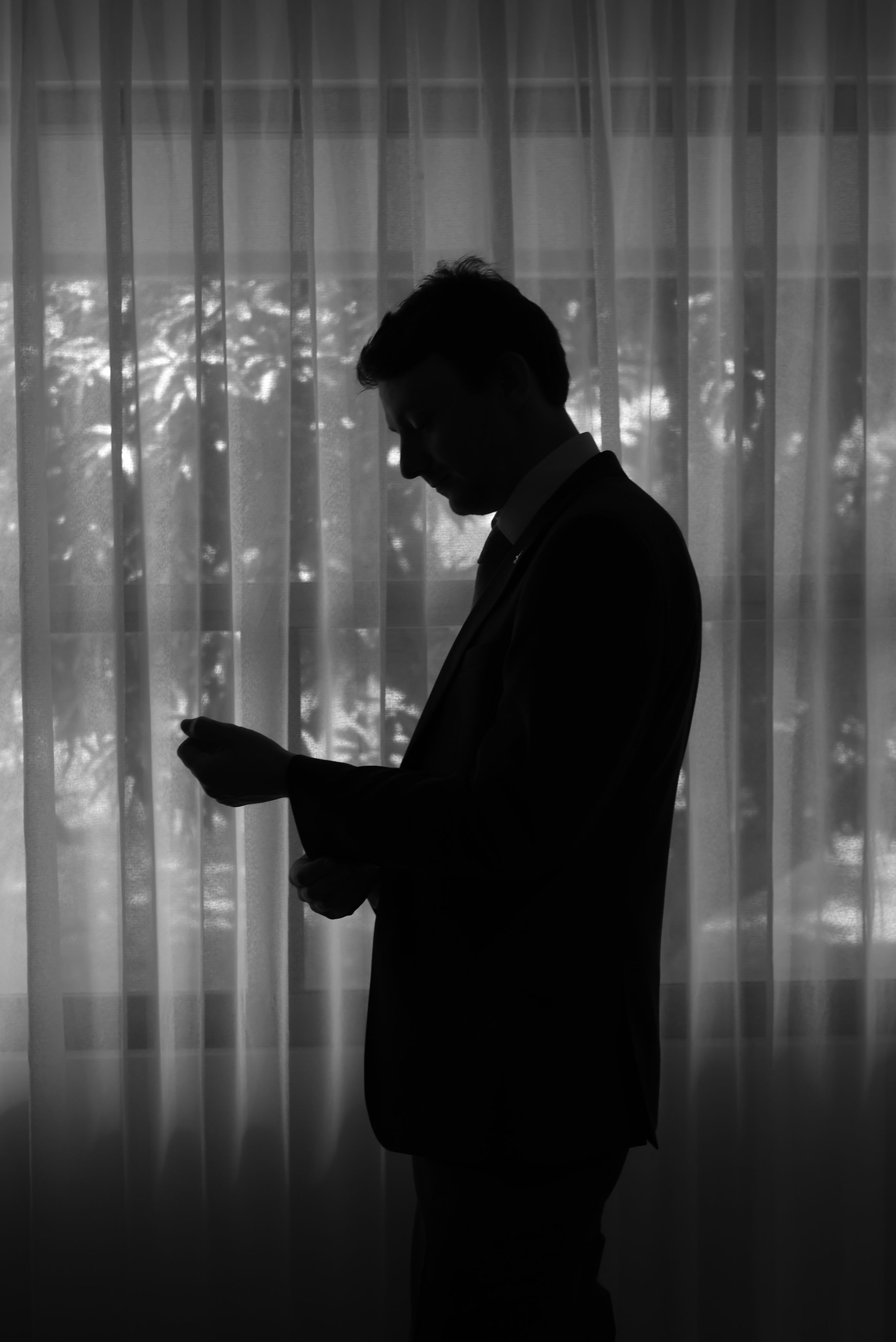 Silhouette of a man in a suit adjusting his cufflinks by a sheer curtained window in black and white.
