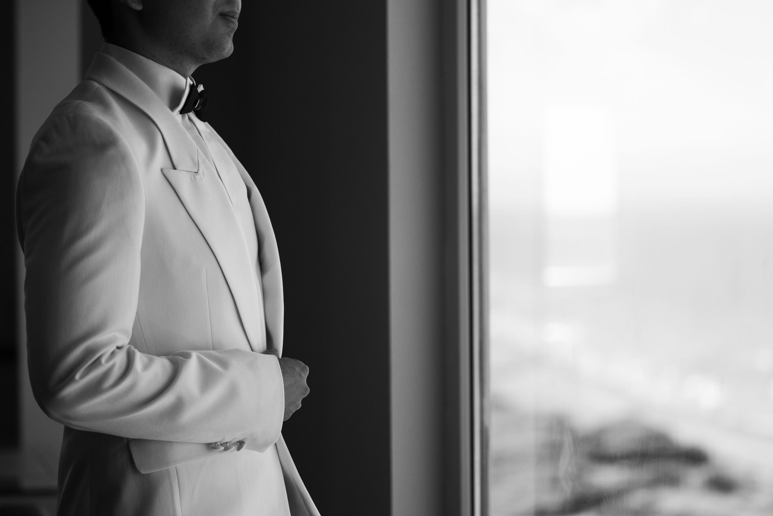 Groom in white tuxedo standing by floor-to-ceiling window with ocean view on wedding day.