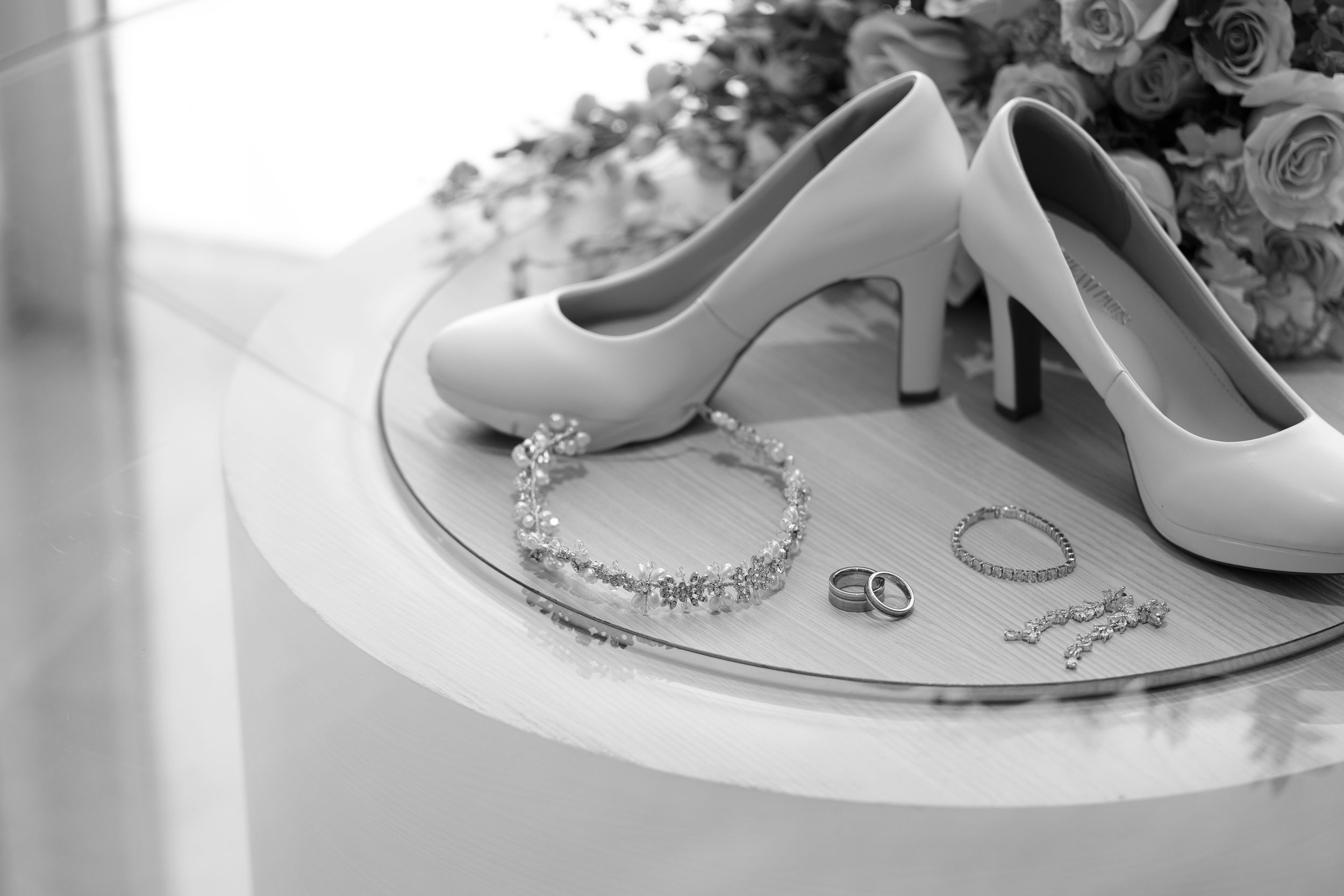 Black and white photo of bridal heels, diamond bracelet, and wedding rings arranged elegantly on a reflective surface.