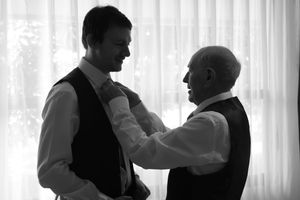 Black and white photo of two men in vests, one adjusting the other's tie near a bright window.