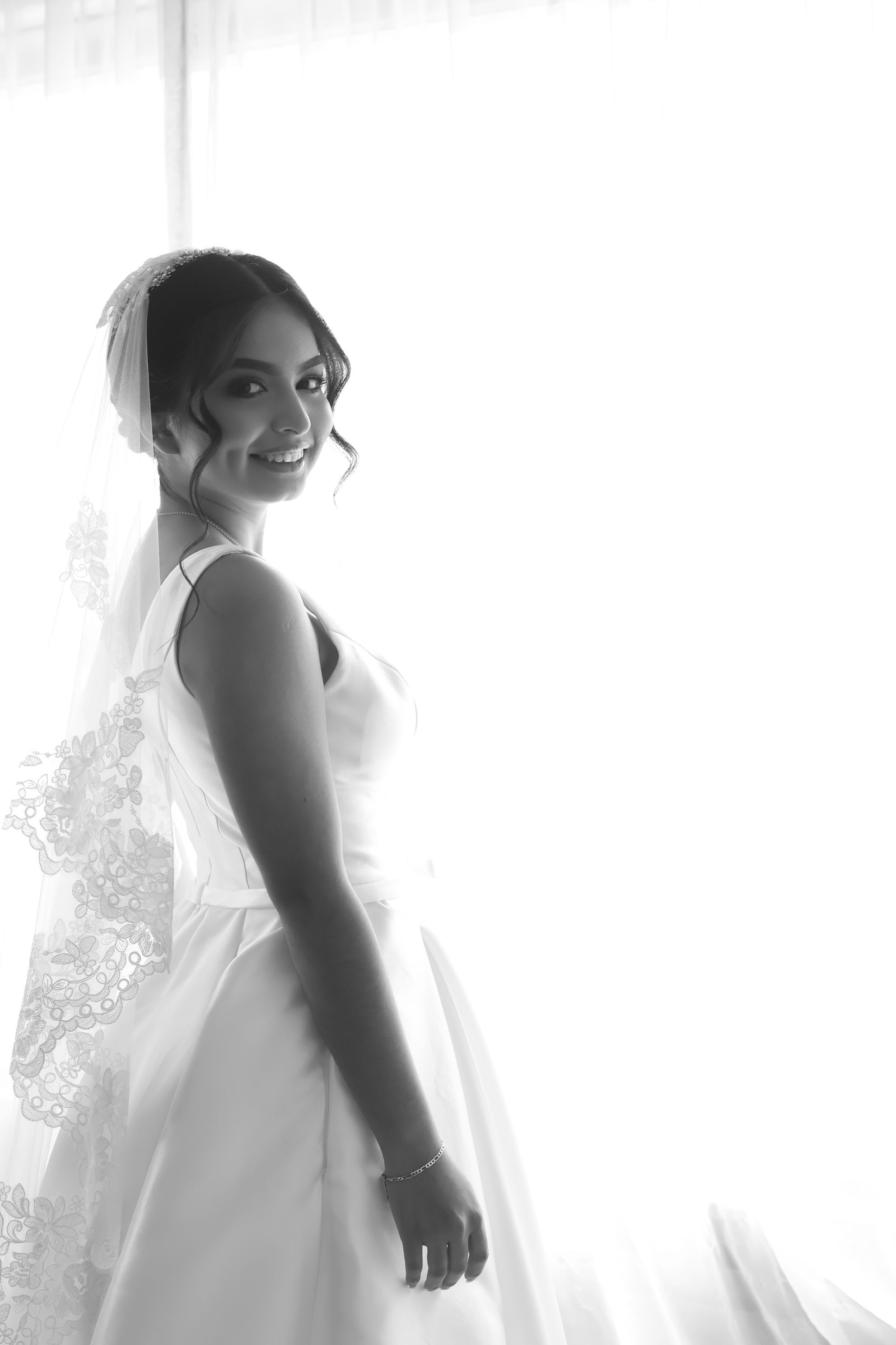 Black and white portrait of a smiling bride in a white gown and veil, standing by a bright window.
