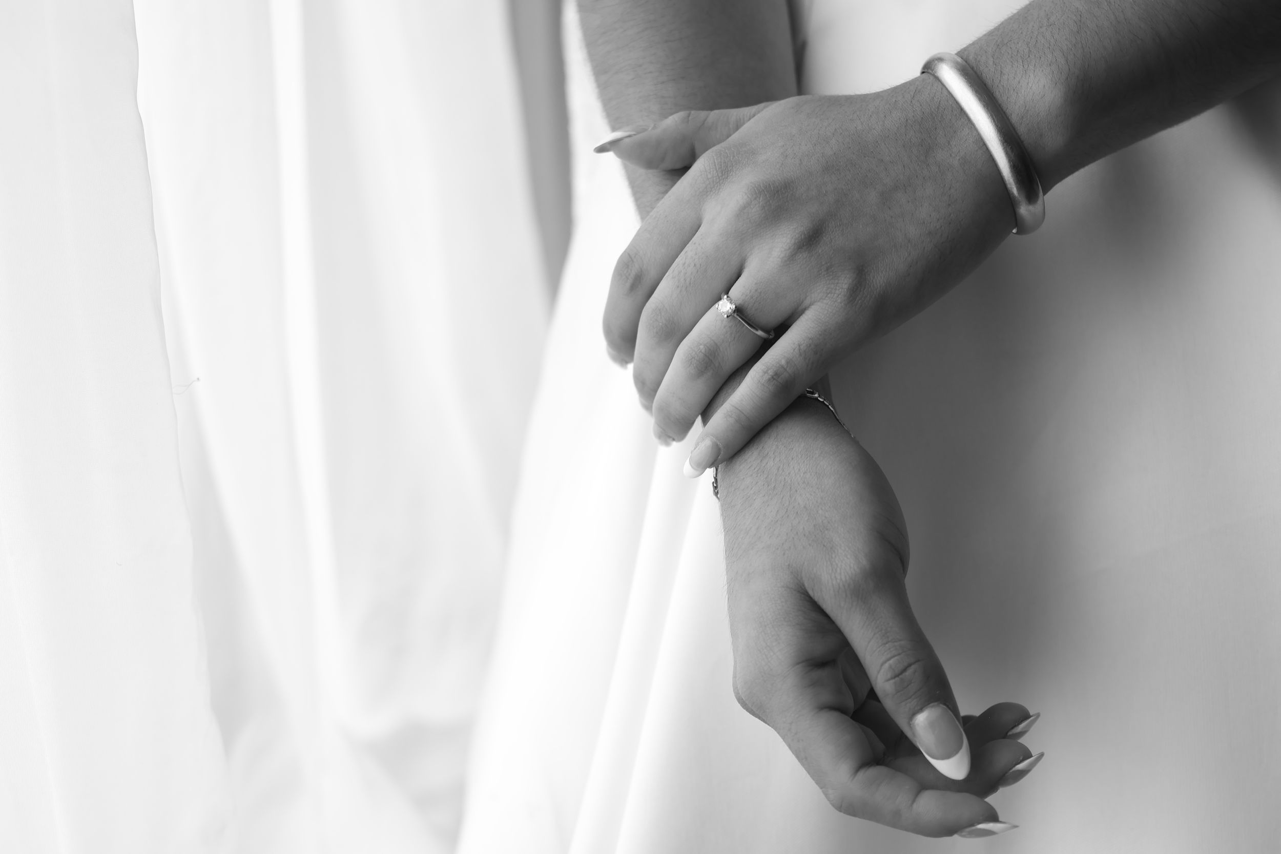Black and white photo of bride's hands with wedding ring and bracelet against white dress.