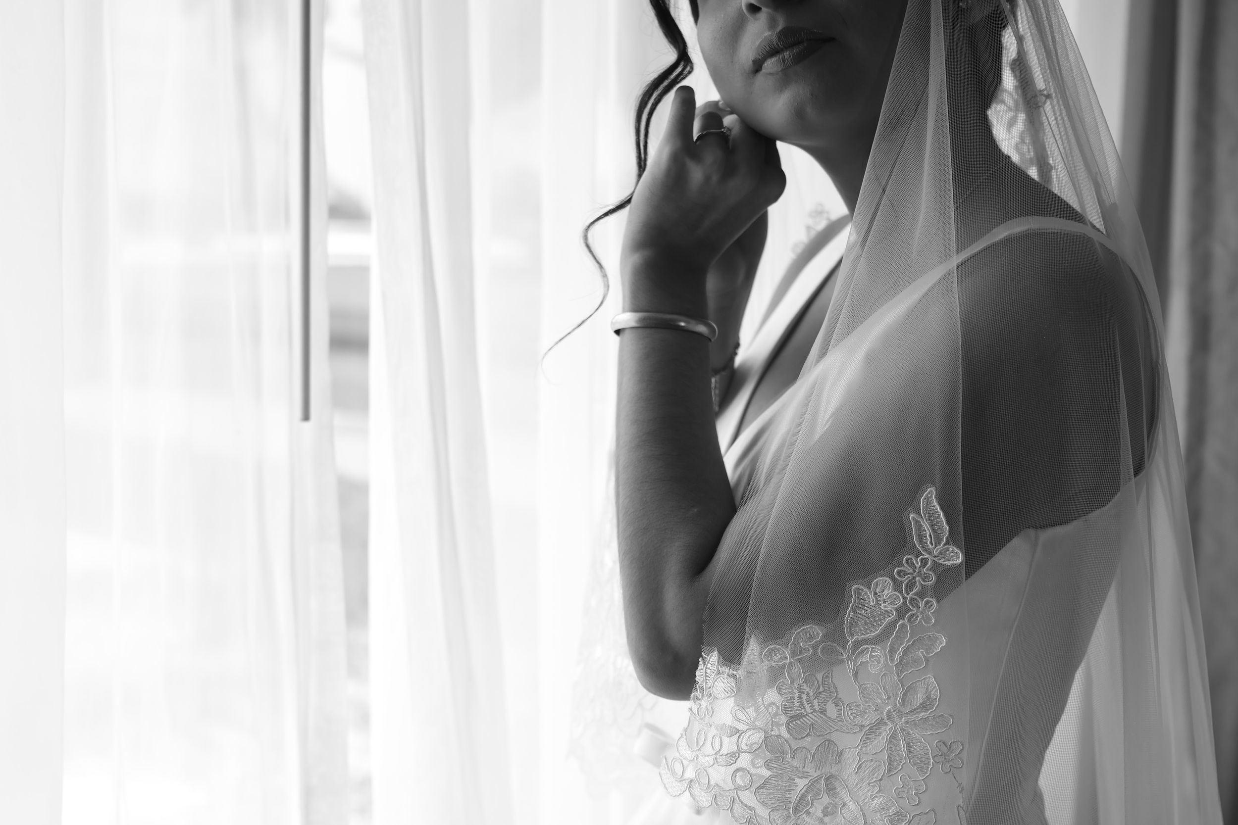 Black and white photo of a bride in lace wedding dress and veil standing by a window in soft natural light.
