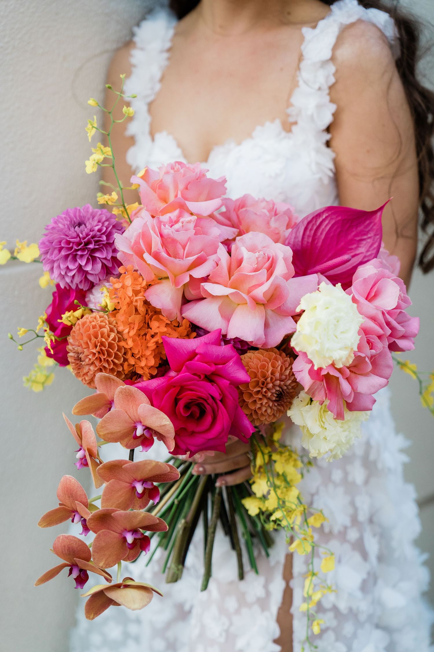 Bride in white lace dress holding vibrant wedding bouquet with pink roses, dahlias, orchids, and yellow blooms.