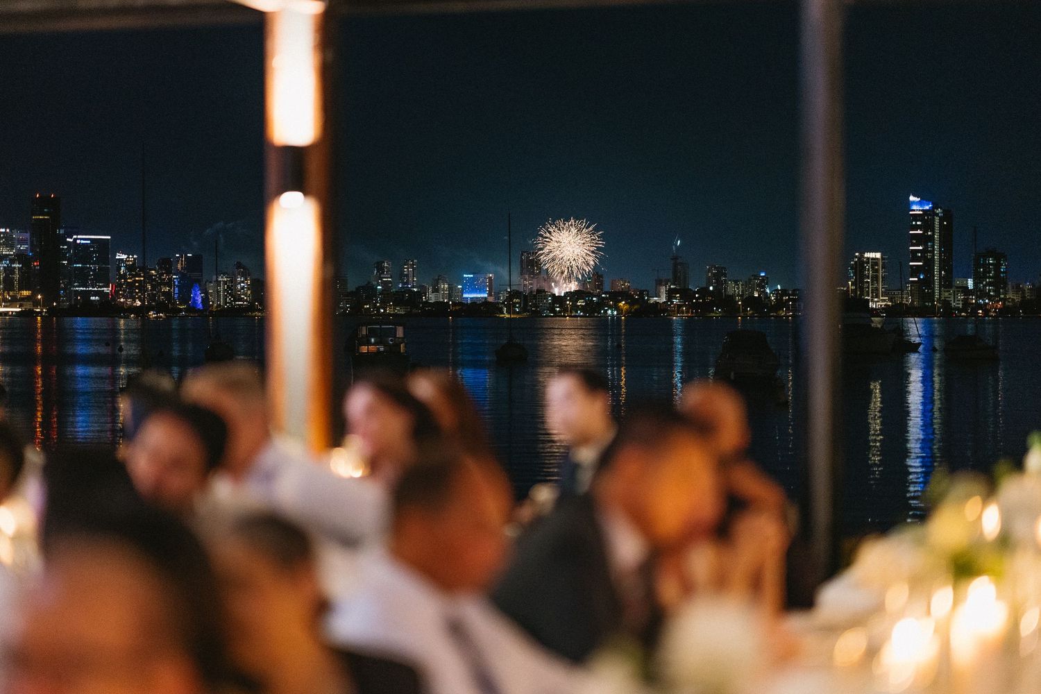 Wedding reception guests seated at tables with a stunning city skyline and fireworks display reflected on the water at night.