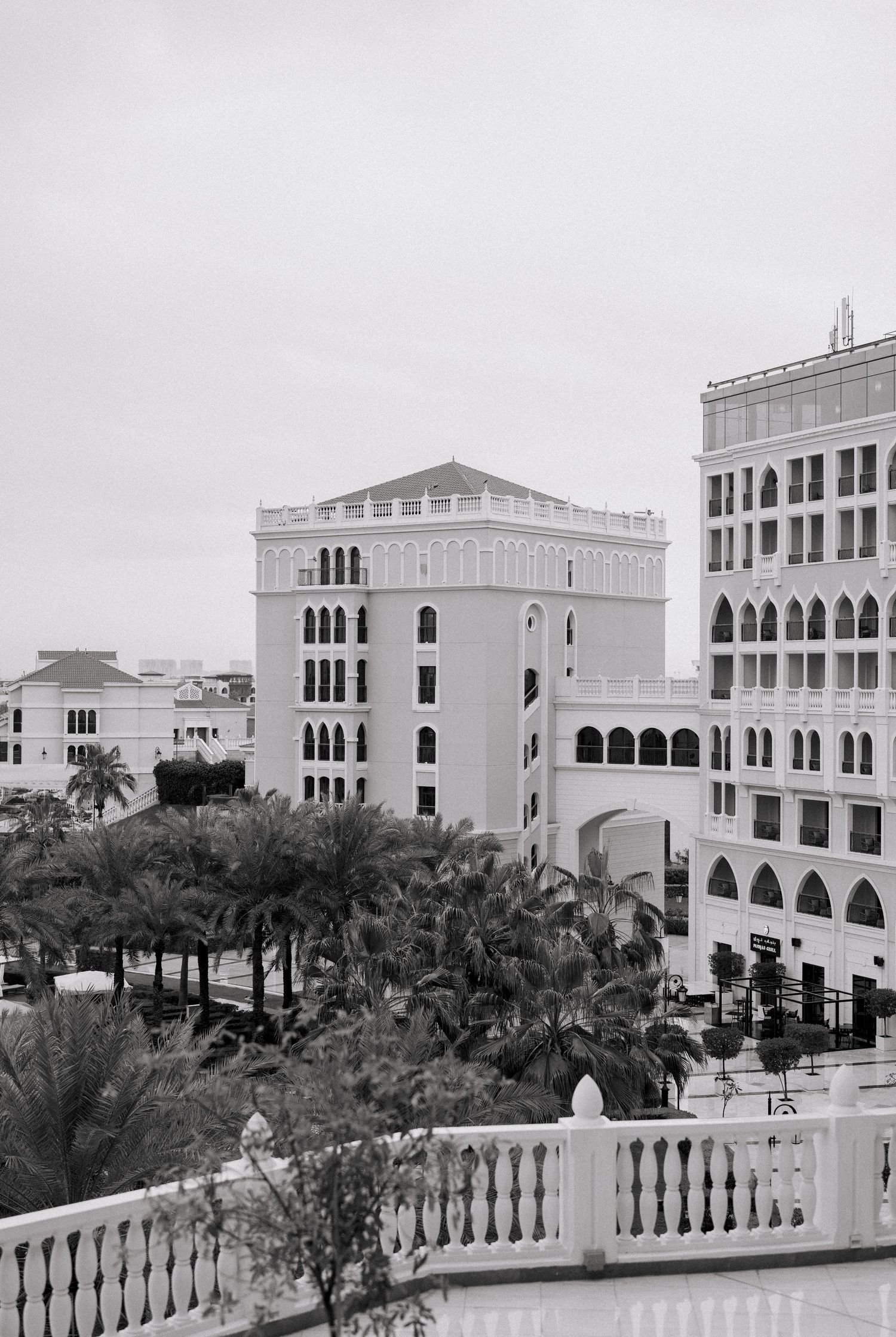 Black and white aerial view of elegant Middle Eastern architecture with arched windows, surrounded by palm trees.