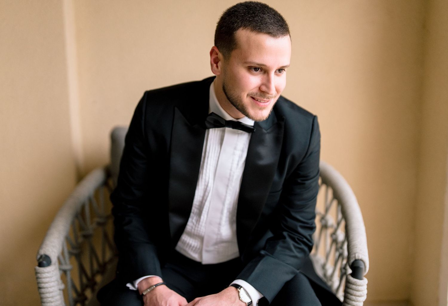 Young man in black tuxedo with bow tie sitting in wicker chair, smiling confidently at formal event.