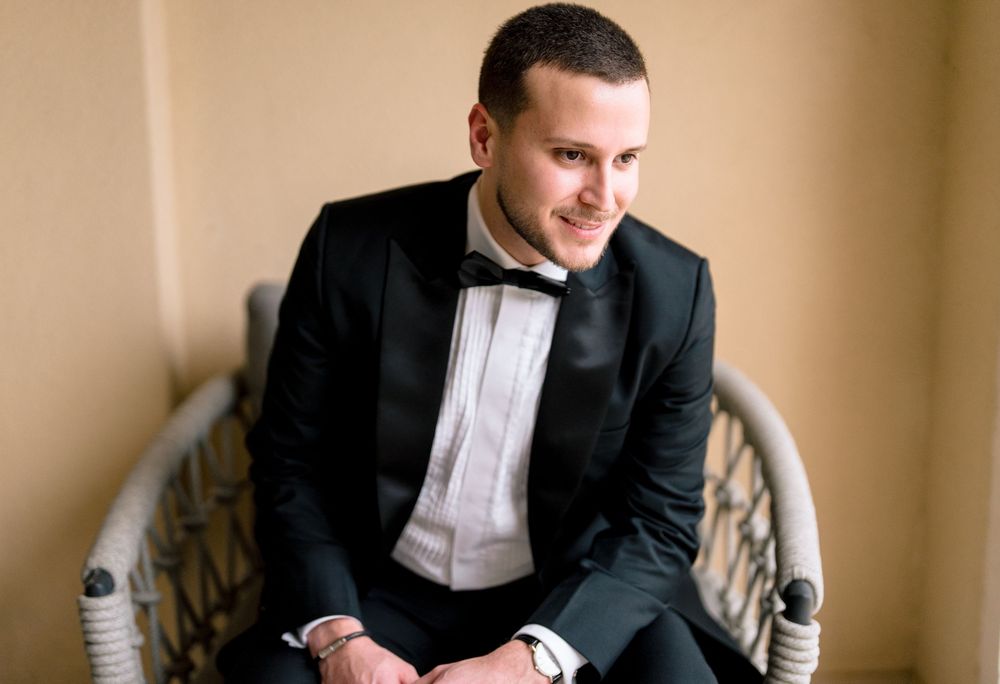 Young man in black tuxedo with bow tie sitting in wicker chair, smiling confidently at formal event.