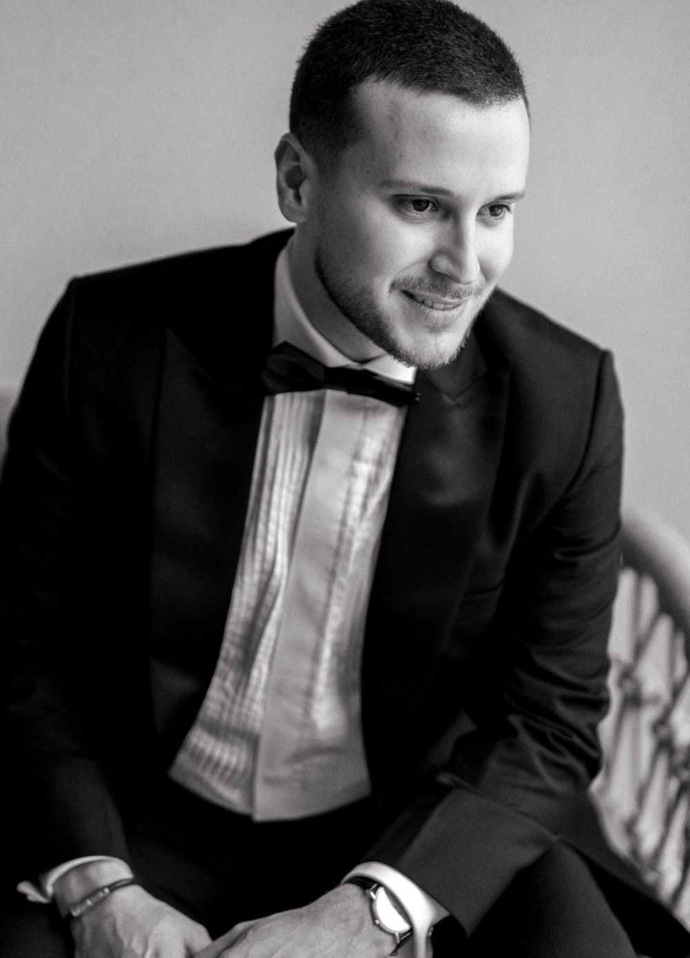 Black and white portrait of a smiling young man in a tuxedo with bow tie, seated in elegant formal attire.