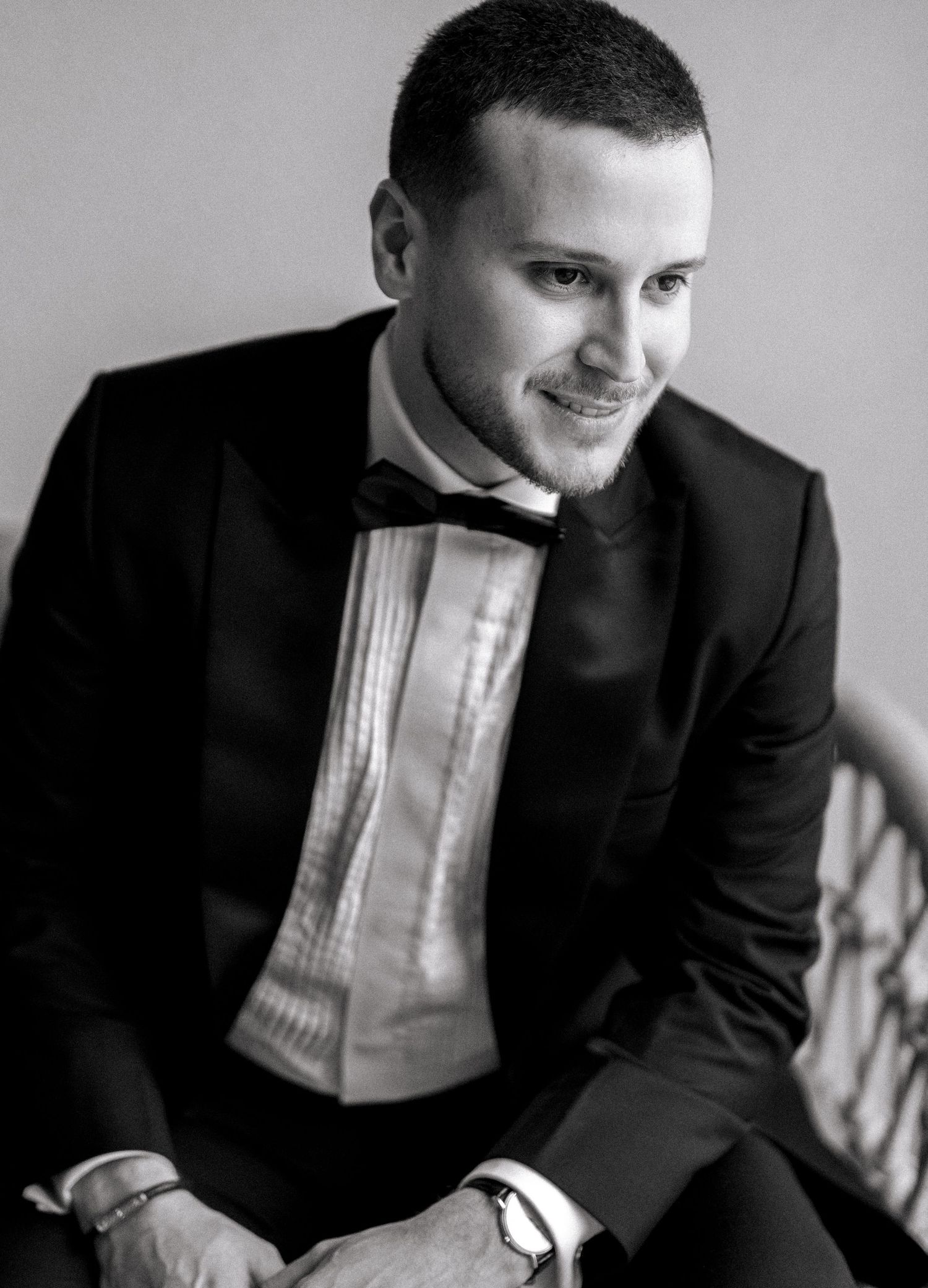 Black and white portrait of a smiling young man in a tuxedo with bow tie, seated in elegant formal attire.