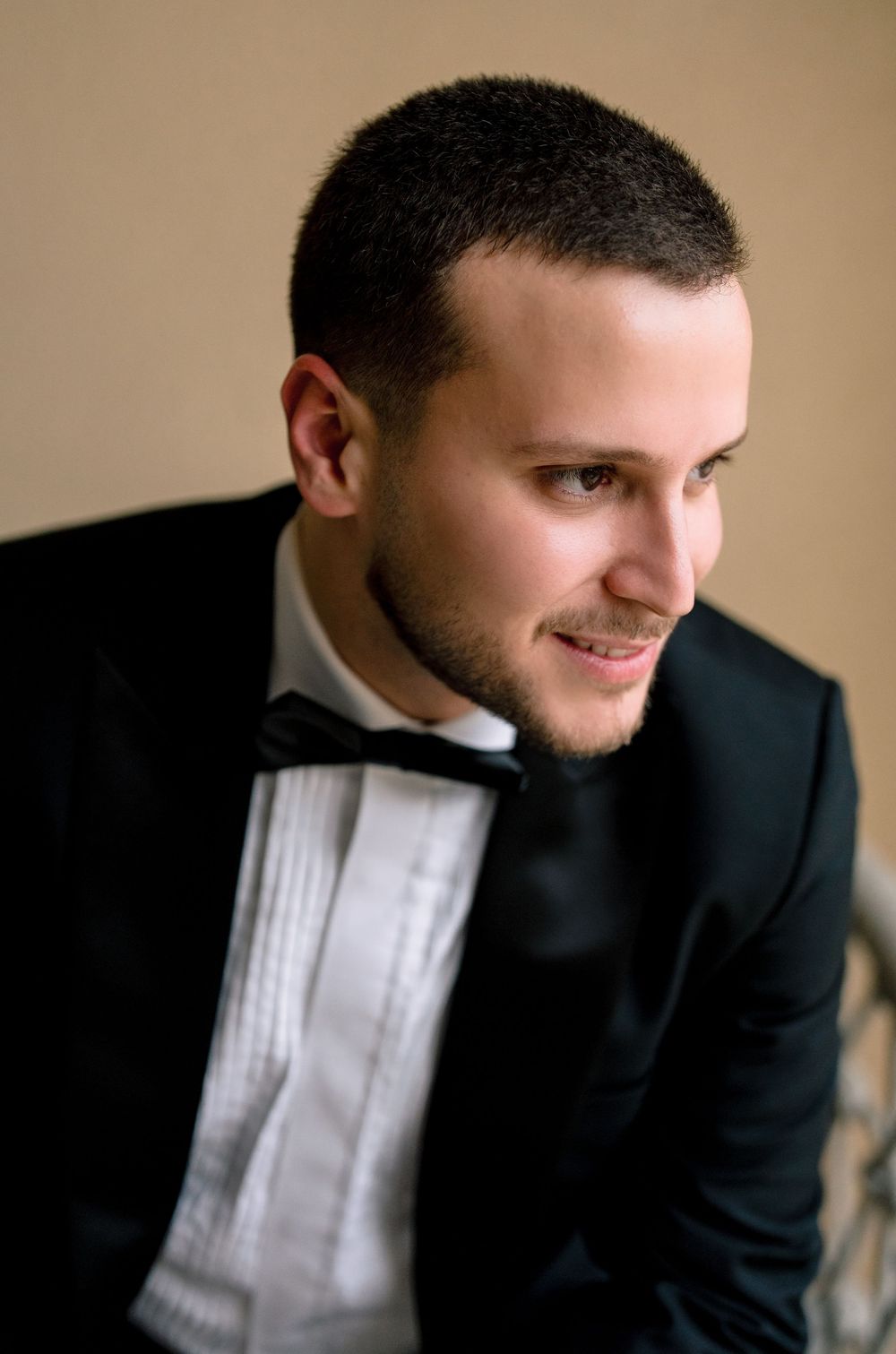 Young man in black tuxedo and bow tie smiling, short dark hair, formal attire portrait photography.