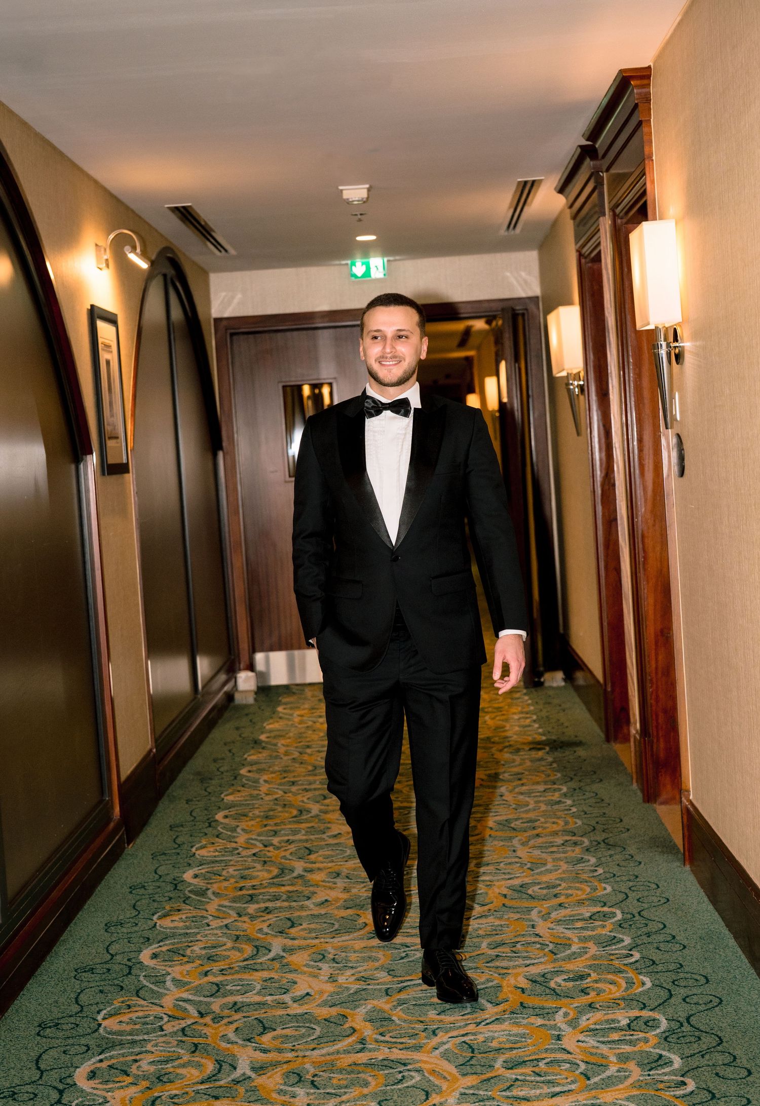 A man in a black tuxedo with bow tie walks confidently down an elegant hotel corridor with green patterned carpet.