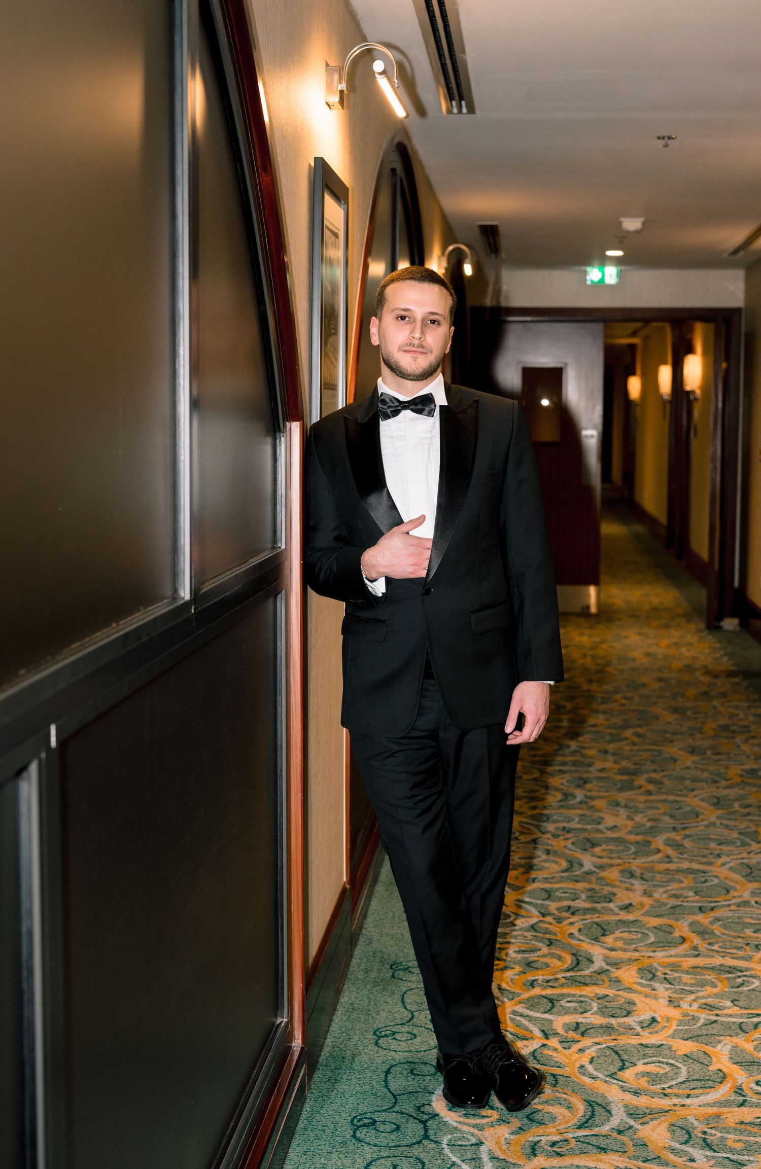 Young man in black tuxedo and bow tie leaning against wall in elegant hotel corridor.