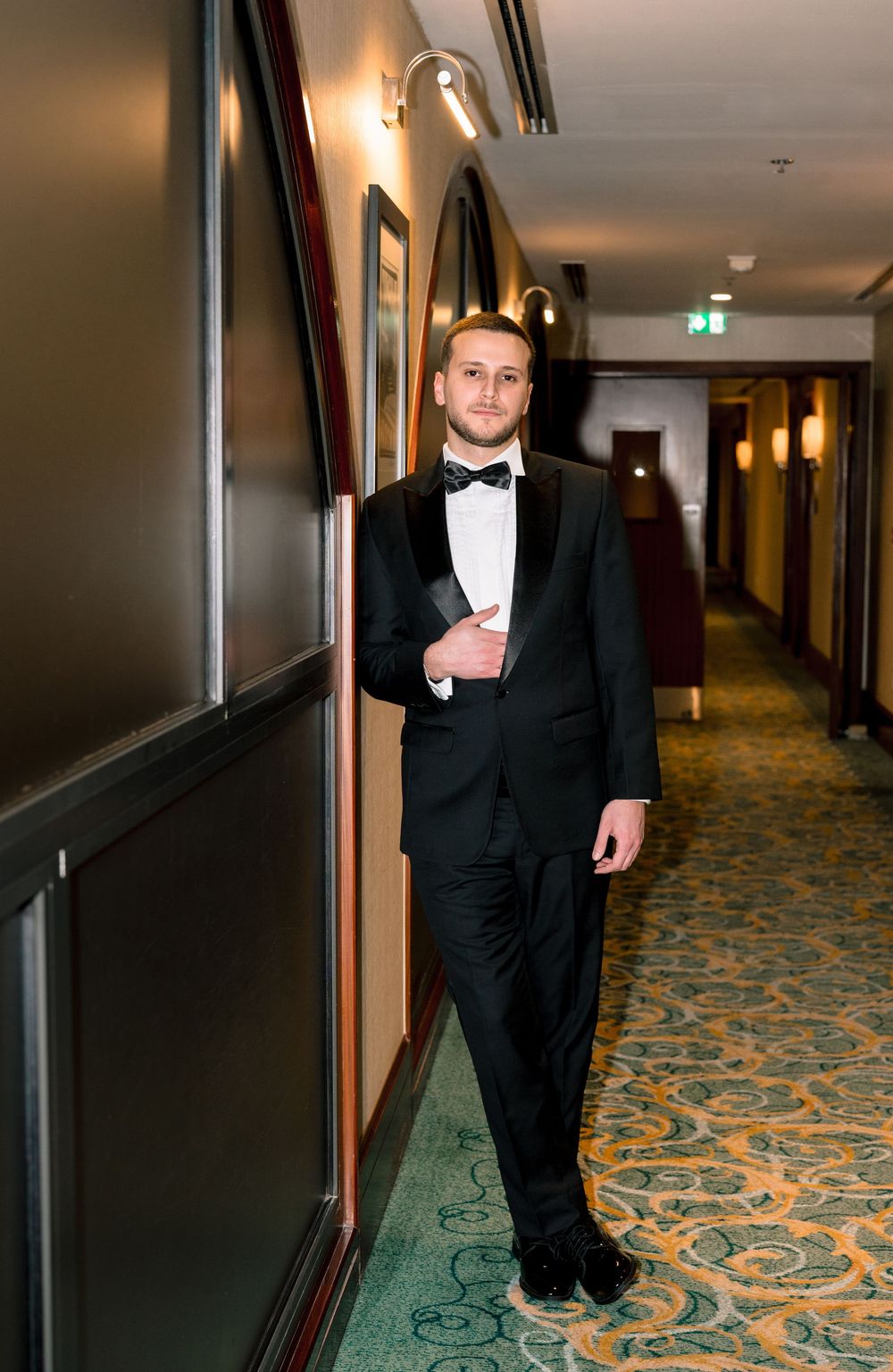 Young man in black tuxedo and bow tie leaning against wall in elegant hotel corridor.