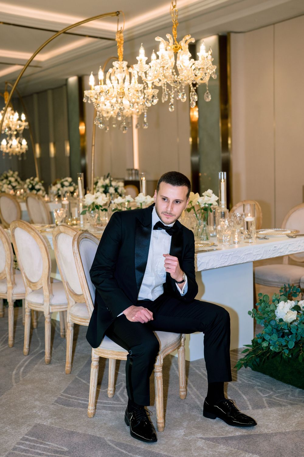 A groom in a black tuxedo sits confidently in an elegant wedding reception hall with crystal chandeliers and floral decor.