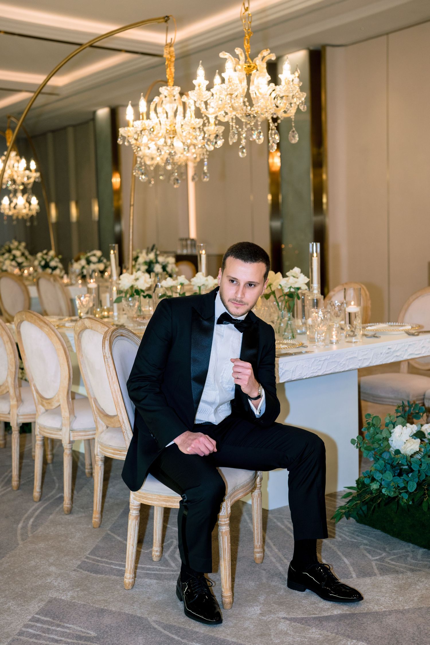 A groom in a black tuxedo sits confidently in an elegant wedding reception hall with crystal chandeliers and floral decor.