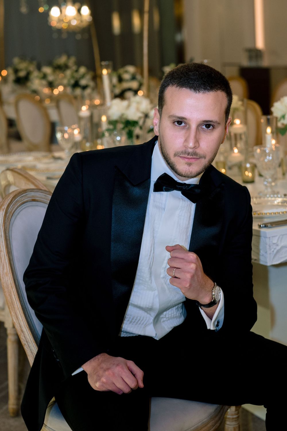 Elegant man in black tuxedo and bow tie seated at a luxurious wedding reception venue.