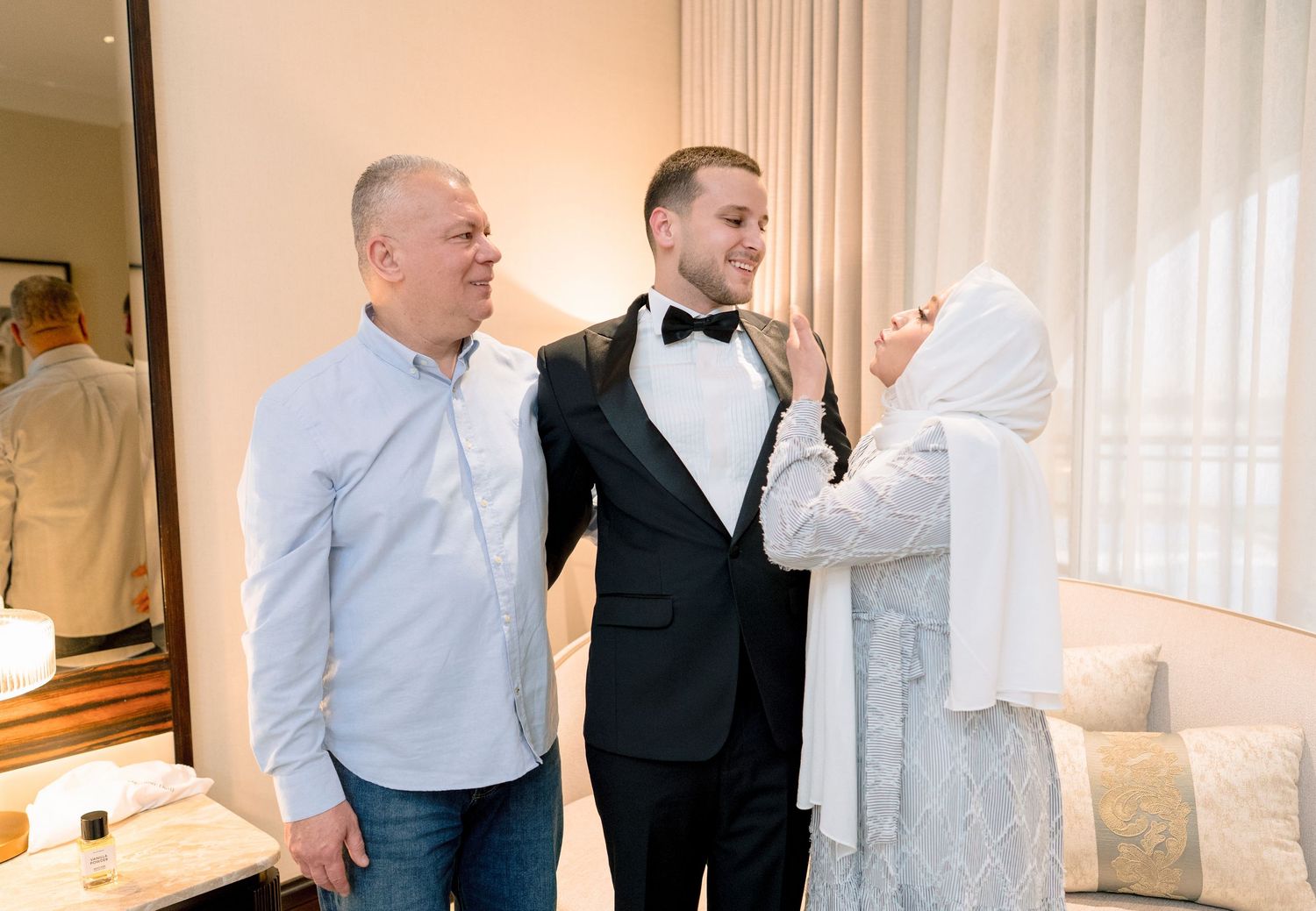 A young groom in a tuxedo stands with his parents as his mother adjusts his bow tie in an elegant room.