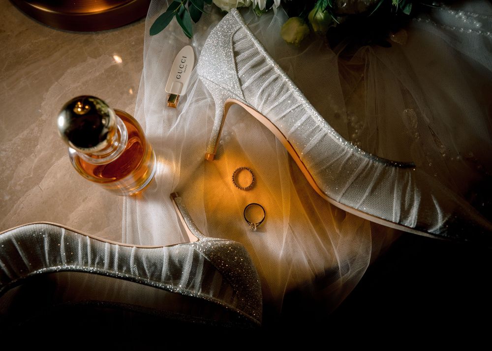 Elegant wedding flat lay with glittery bridal shoes, gold rings, perfume bottle, and veil on a warm golden surface.