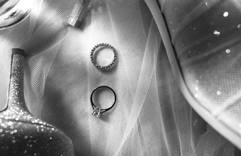 Black and white photo of wedding rings and diamond band resting on bridal veil with glittery heels nearby.