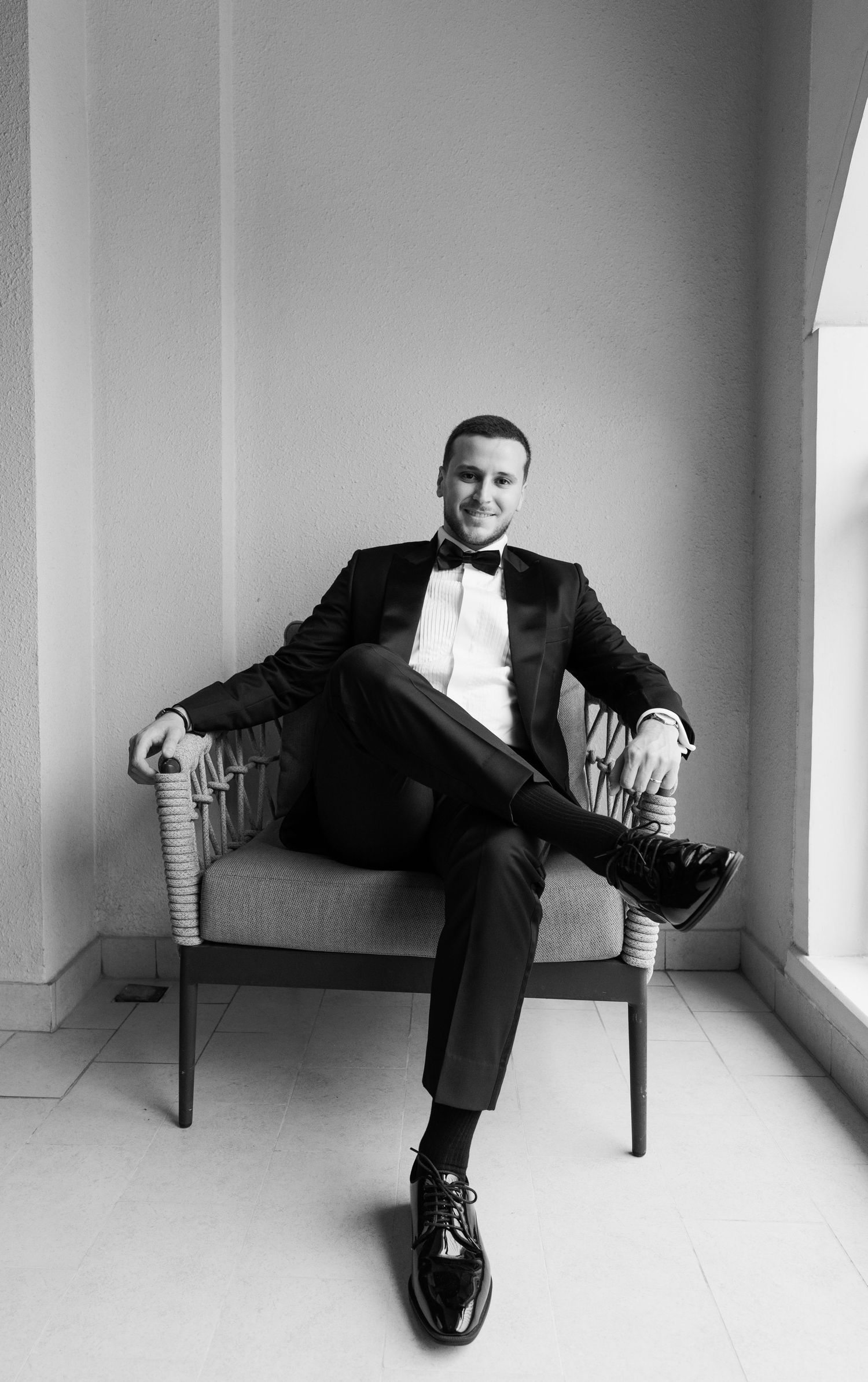 Man in black tuxedo and bow tie sitting relaxed in chair, black and white formal portrait photo.