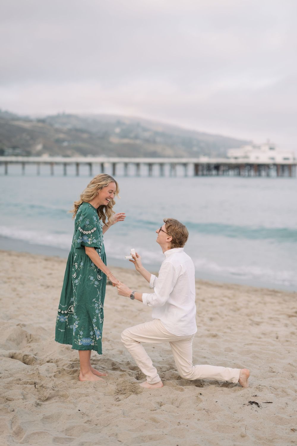 Man proposes to woman on sandy beach near pier, she reacts with joy wearing green floral dress.