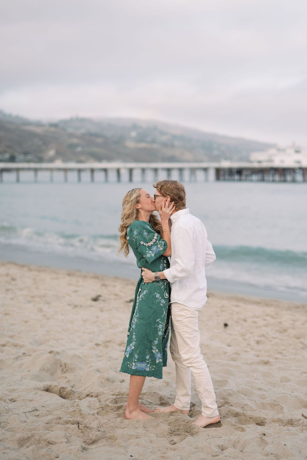 Couple kissing barefoot on sandy beach near pier, woman in teal dress, man in white outfit, overcast sky.