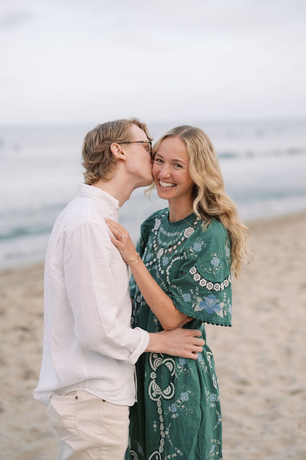 Couple sharing a romantic kiss on a sandy beach, woman in green floral dress smiling joyfully by the ocean.